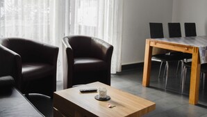 Ein modernes Wohnzimmer mit einem Holztisch, zwei Ledersesseln, einem Esstisch mit vier Stühlen und einem großen Fenster mit transparenten Vorhängen.