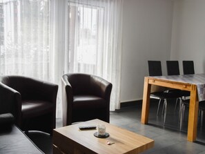 Ein modernes Wohnzimmer mit einem Holztisch, zwei Ledersesseln, einem Esstisch mit vier Stühlen und einem großen Fenster mit transparenten Vorhängen.