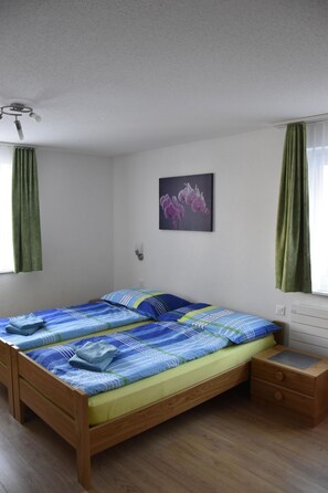 Ein Doppelbett mit blau-grün gestreifter Bettdecke, ein Nachttisch aus Holz mit Schubladen, eine Wandleuchte und ein Fenster mit grünen Vorhängen.