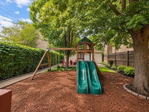 A playground with a wooden playhouse and slides, surrounded by a mulched area and a hedge.