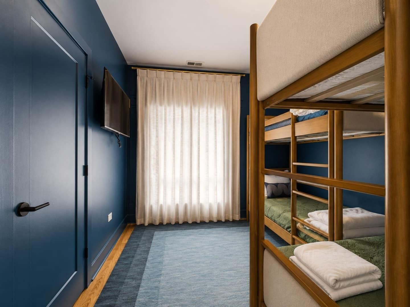 A room with a bunk bed, a door, a flat-screen TV, and a window with curtains.