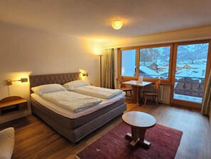 Et hotelværelse med en seng, et lille bord, en stol, et vindue med udsigt over et sneklædt landskab og en rød måtte.