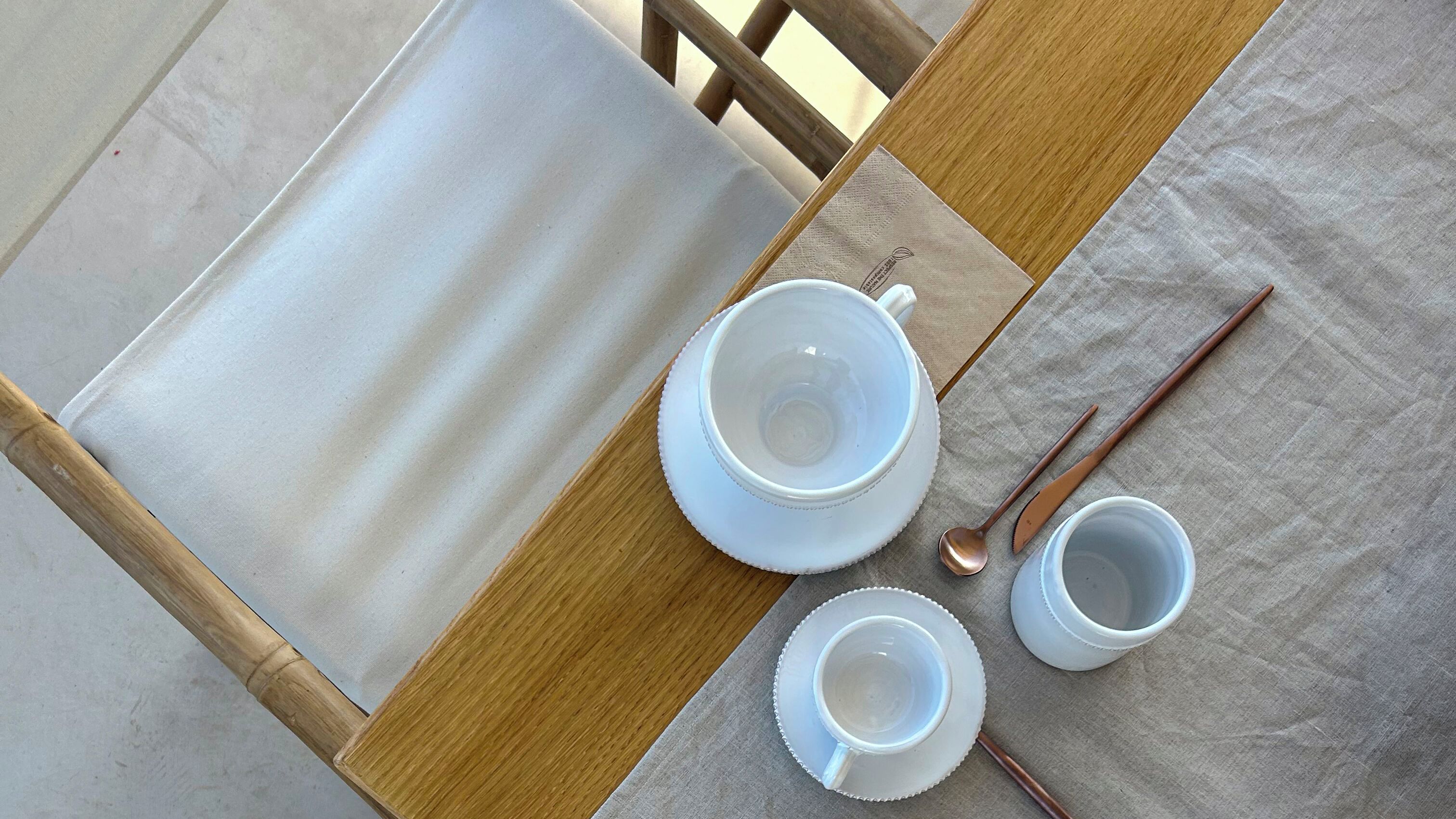Taula amb un plat blanc, una tassa blanca, una mug blanca i una cullera sobre un mantell beige.