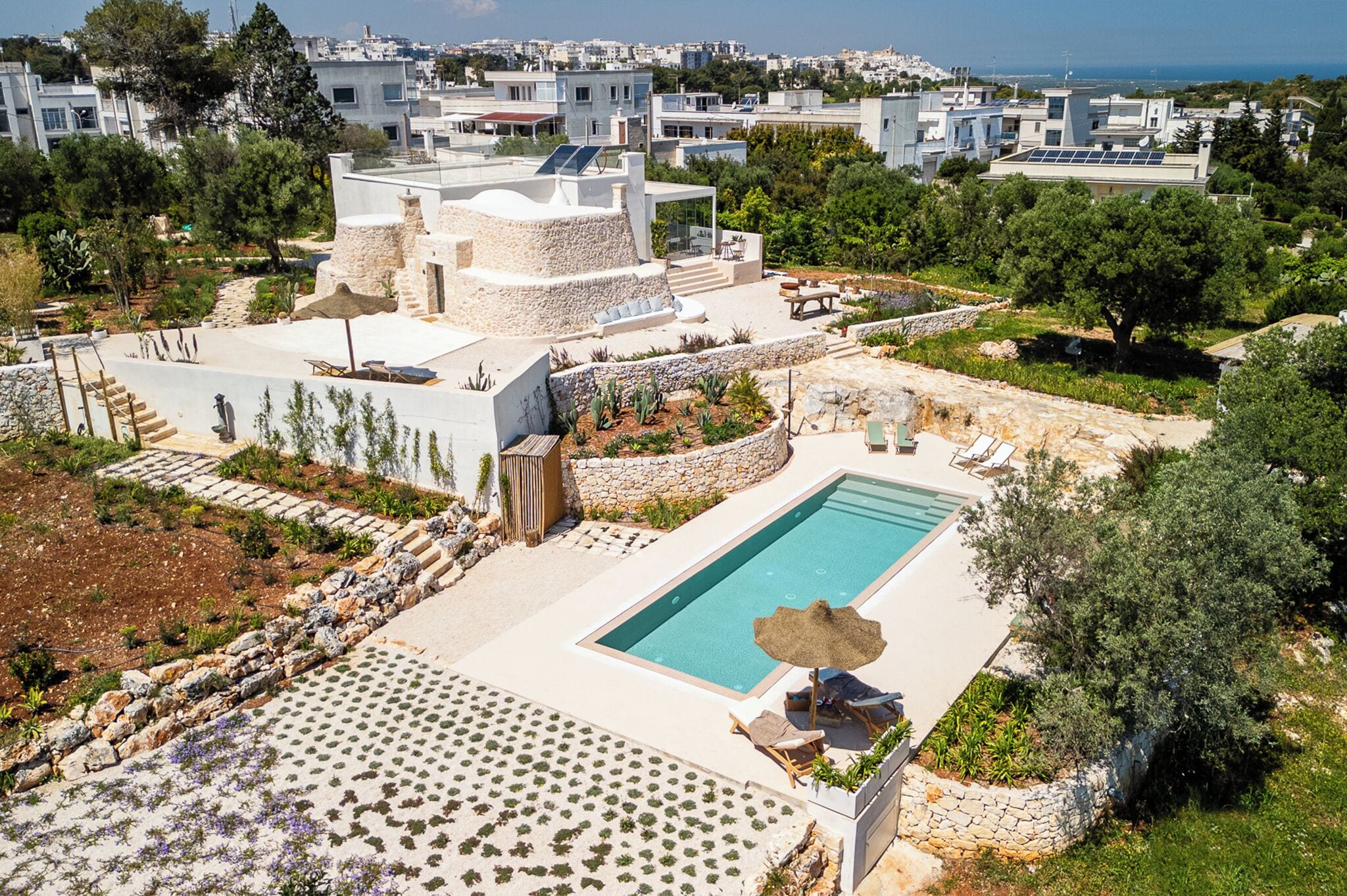 A rooftop pool area with a view of the cityscape, surrounded by greenery and stone structures.