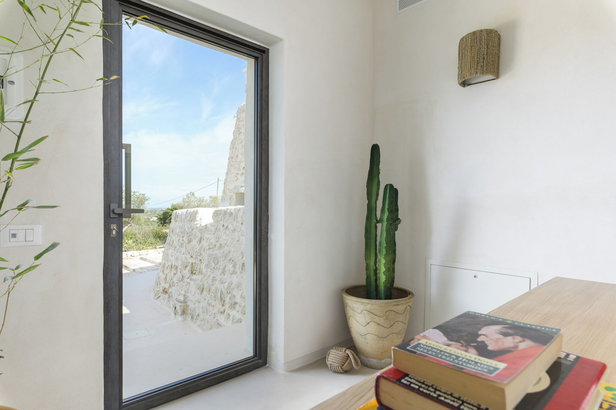 Una habitación con puerta de vidrio corredera que da a un espacio al aire libre, un cactus en maceta y una pila de libros sobre una mesa.