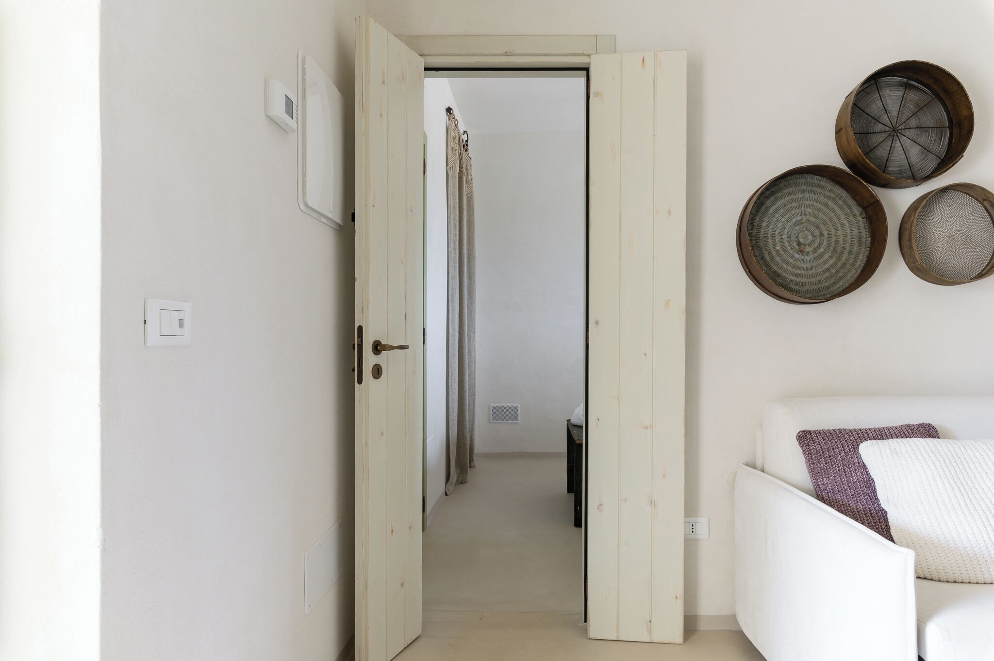 Un couloir avec une porte blanche, un mur blanc et un canapé blanc agrémenté d’un coussin à motifs. Trois paniers en osier sont accrochés au mur.