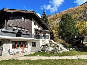 Un chalet de dos pisos con balcón, cimientos de piedra y vistas a un paisaje montañoso.