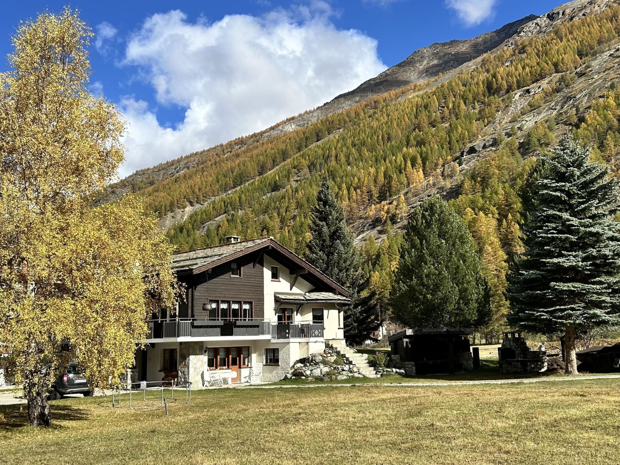 Un chalet à deux étages doté d’un balcon, entouré d’une pelouse et d’arbres, avec une chaîne de montagnes en toile de fond.