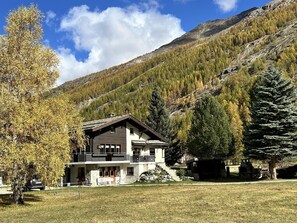 Un chalet de dos pisos con balcón, rodeado de césped y árboles, con montañas al fondo.