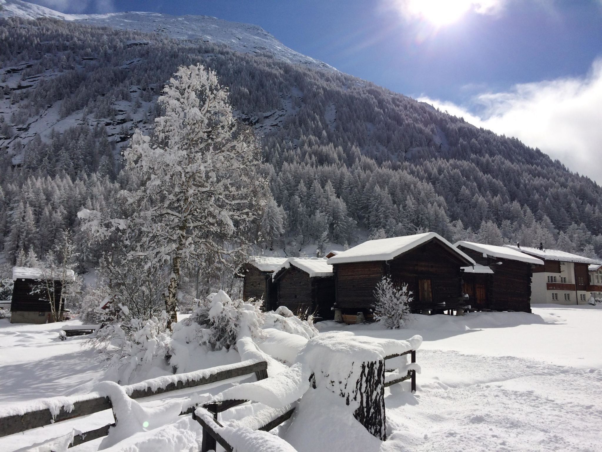 Un chalet enneigé avec des cabanes en bois, entouré d’une forêt et d’une montagne.