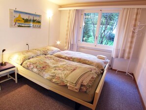 Cama doble con ropa de cama floral, una mesita de noche con lámpara, una ventana con cortinas y un cuadro colgado en la pared.