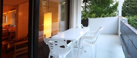 Un balcon extérieur avec une table et des chaises blanches, une porte vitrée donnant sur un espace intérieur et une vue sur la verdure.
