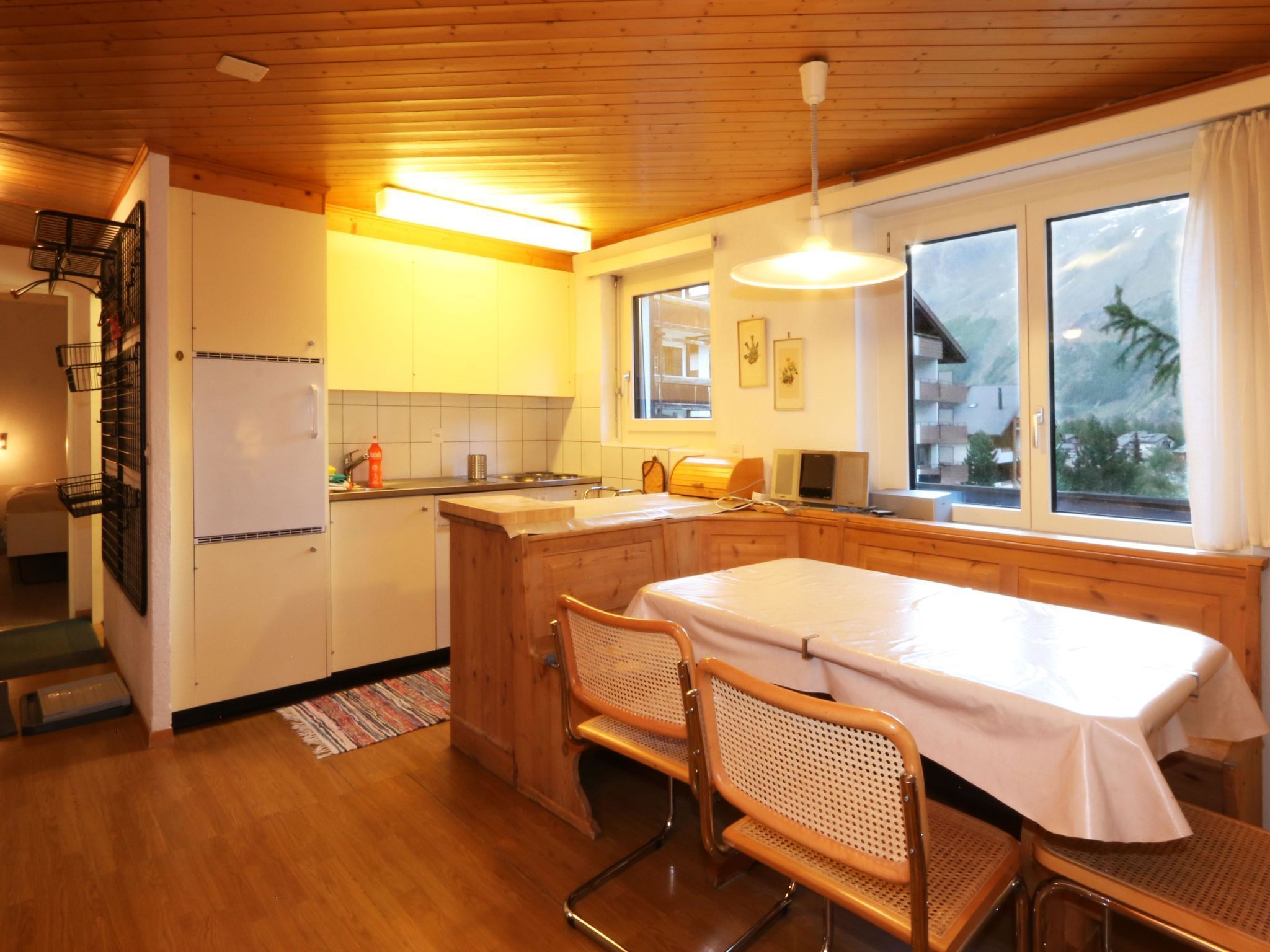 Een keuken met houten kastjes, een eettafel met stoelen, een koelkast en een raam met uitzicht op een berg.