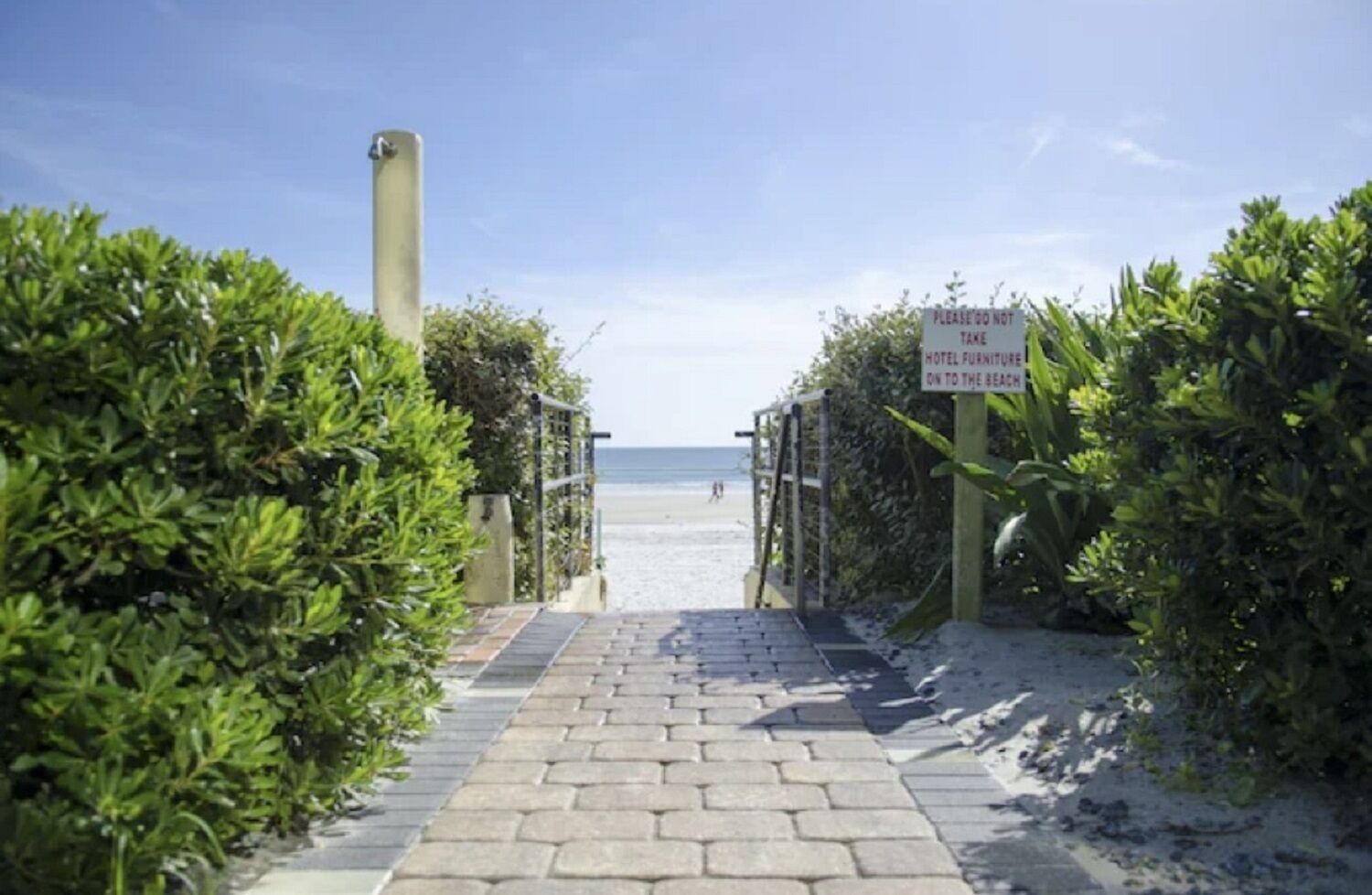 Ein gepflasterter Weg führt zu einem Strand, auf dem ein Schild das Tragen von Hotelmöbeln zum Strand verbietet.