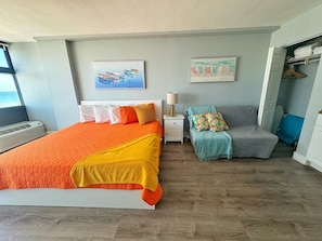 Een slaapkamer met een groot bed, een nachtkastje met een lamp, een grijze bank en twee wanddecoraties.