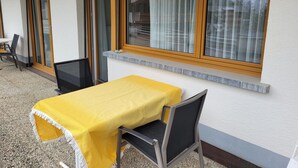 Un patio con tavolo coperto da una tovaglia gialla, due sedie e una finestra con tende.
