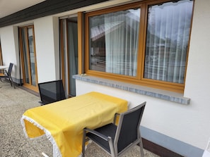A patio with a table covered by a yellow cloth, two chairs, and a window with curtains. - Haus Perle Appartement/Fewo 3-Bettwohnung Nr. 2 by Interhome (Saas-Fee)
