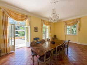 Een eetkamer met een grote houten tafel, stoelen, een kroonluchter en gordijnen.