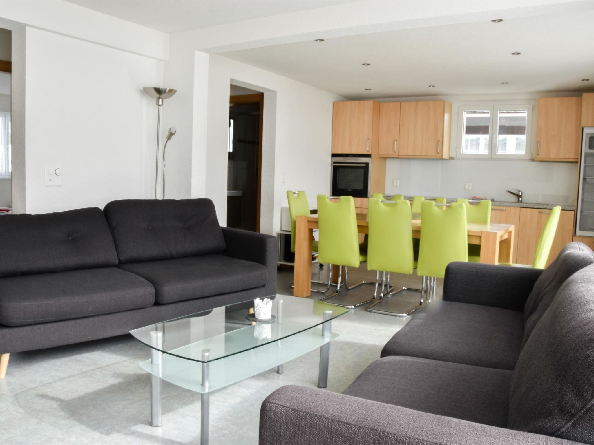 A modern living room with a glass coffee table, two grey sofas, and a dining area with green chairs.