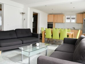 Ein modernes Wohnzimmer mit einem Glastisch, zwei grauen Sofas und einem Essbereich mit grünen Stühlen.