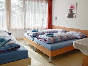 A hotel room with two beds, a wooden headboard, a window with sheer curtains, a painting on the wall, and a white sink. - Heidi Appartement/Fewo 5-Bettwohnung Nr. 63 by Interhome (Saas-Fee)