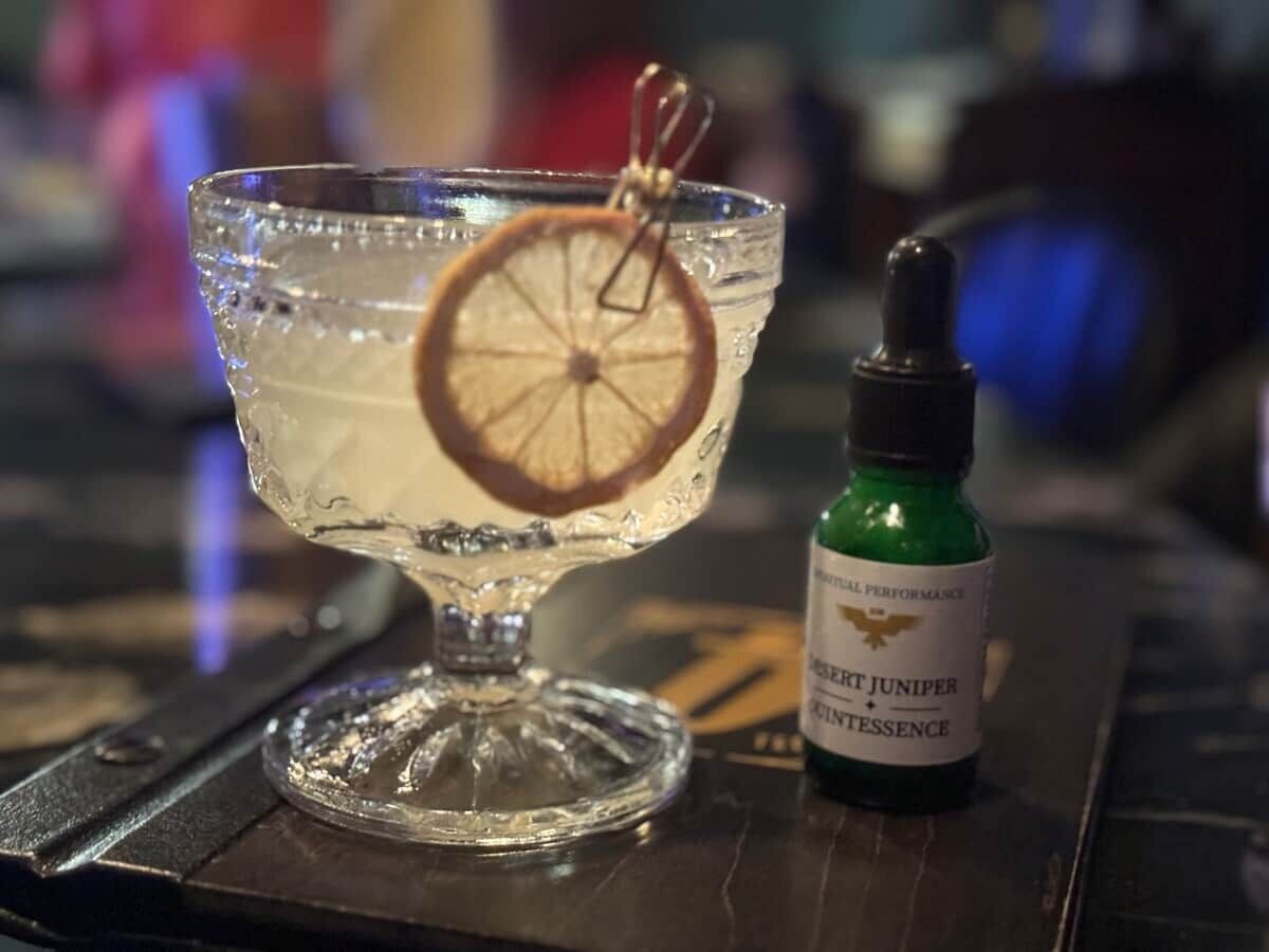 Un verre transparent contenant un cocktail, garni d’une tranche de citron, et un flacon compte-gouttes étiqueté « JUNIPER » posé sur le comptoir d’un bar.