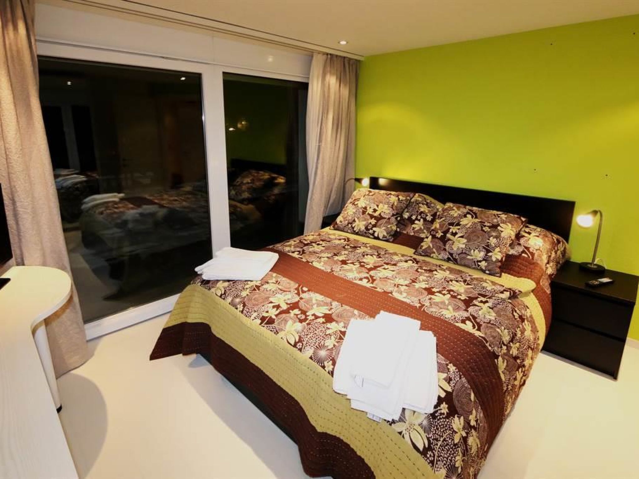 Une chambre à coucher avec un grand lit, une table de chevet, une lampe et une vue sur l’extérieur, accessible par une porte-fenêtre coulissante.
