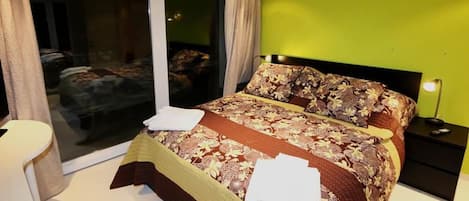 Un dormitorio con una cama grande, una mesita de noche, una lámpara y vistas a través de un ventanal corredero.