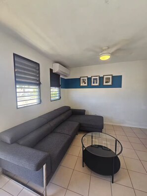 A living room with a sectional sofa, a glass coffee table, and a wall-mounted air conditioner. - Spacious 3BR steps to Beach- Family friendly (San Juan)