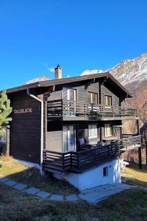 Ein zweistöckiges Holzhaus mit Balkon, beschriftet mit 'TALBLICK', vor einer Bergkulisse.