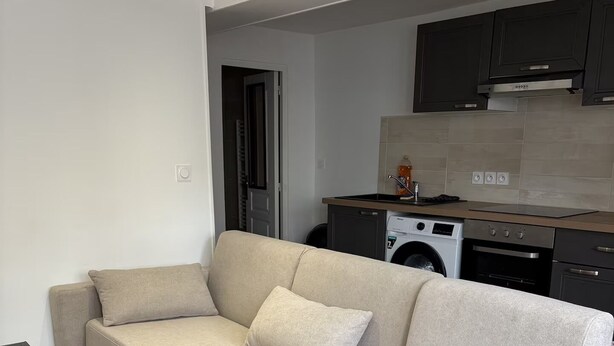 Ein modernes Wohnzimmer mit einem beigefarbenen Ecksofa, einem kleinen schwarzen Beistelltisch und einem Küchenbereich mit Waschmaschine, Backofen und Schränken.