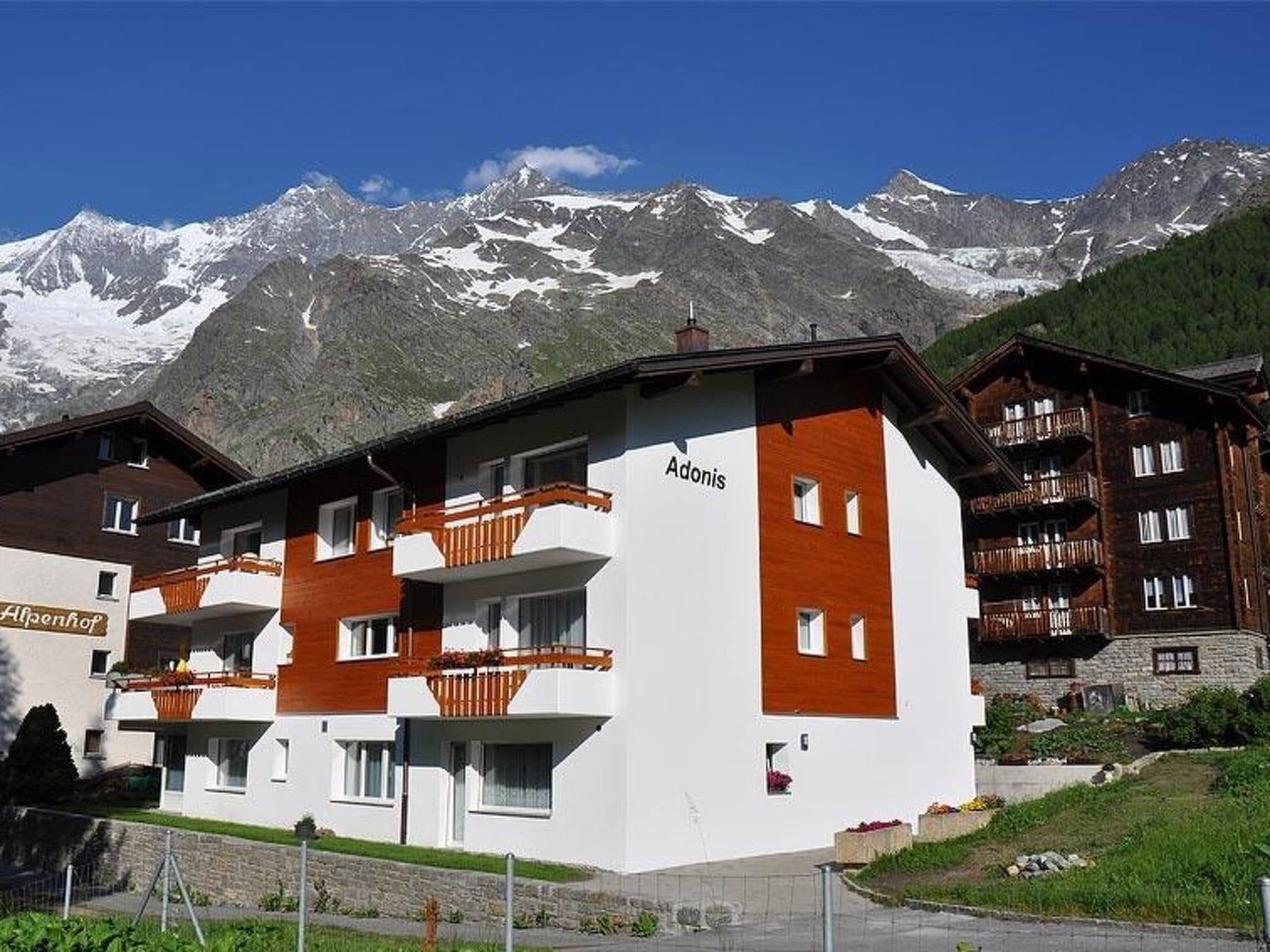 Un hotel de varios pisos con balcones, llamado 'Adonis', con montañas nevadas al fondo.