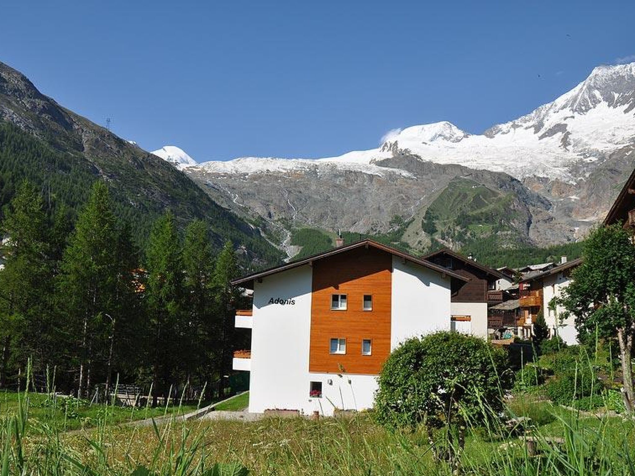 Un edificio de dos pisos con fachada blanca y detalles de madera, rodeado por un paisaje montañoso con picos nevados.
