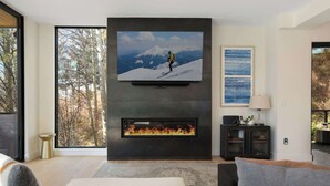 Una sala de estar moderna con chimenea, un televisor de pantalla plana grande montado en la pared y vistas a árboles a través de ventanas amplias.