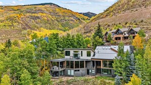 Une maison moderne à deux étages, dotée de grandes fenêtres et d’un balcon, entourée d’une forêt dense et d’un feuillage automnal.