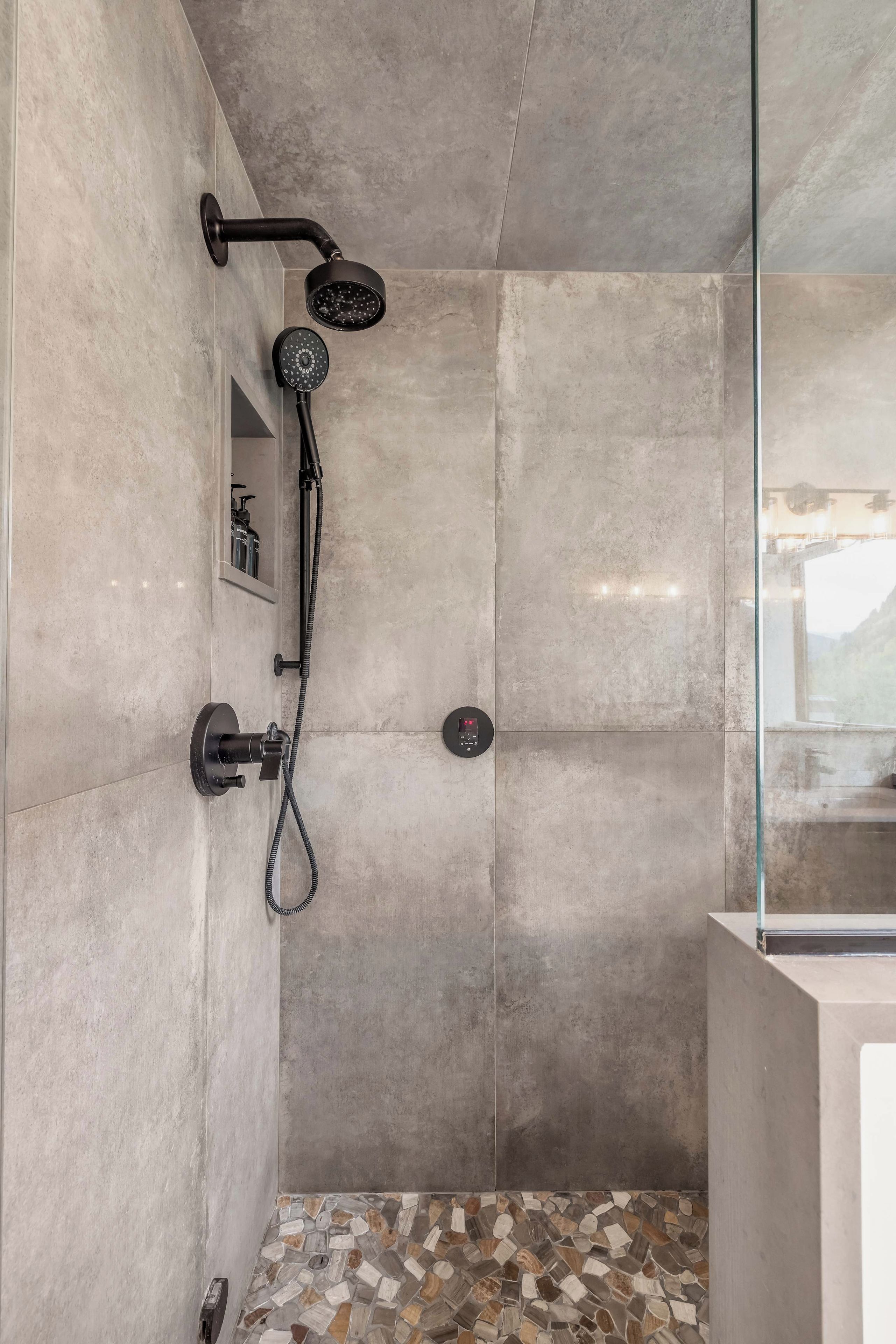 A modern shower with a handheld showerhead and a fixed showerhead, a control panel, and a tiled floor with a mosaic pattern.
