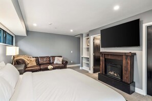 A bedroom with a large bed, a TV mounted on the wall, a fireplace, and a window with blinds. - East Vail Retreat | Hot Tub, Fire Pit & Views (Vail)