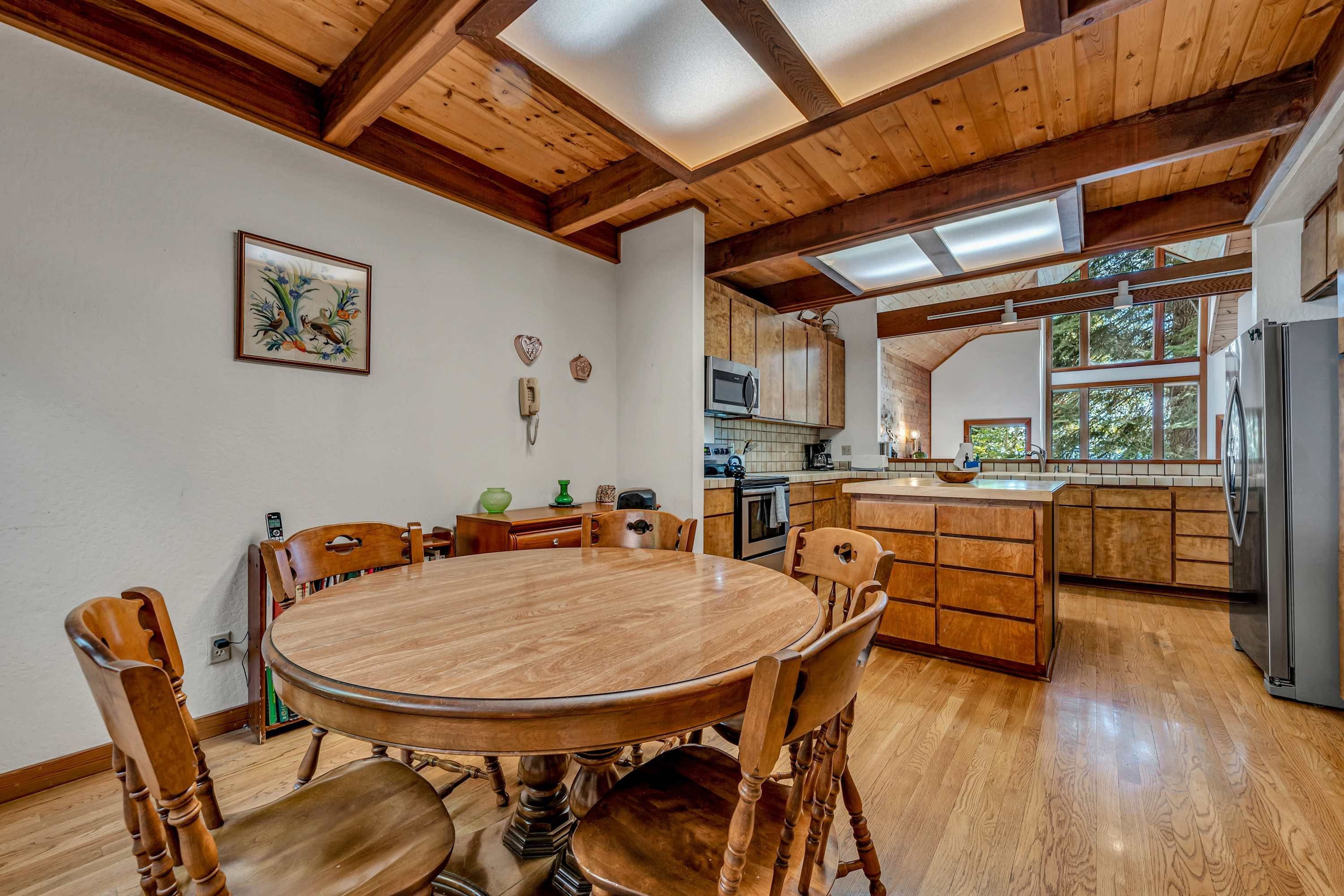 A wooden dining table with chairs, a kitchen with wooden cabinets, and a large window.