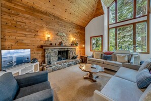 A cozy living room with a stone fireplace, wooden walls, and a large window overlooking greenery. - Tahoe Waterfront Oasis | Deck & Lake Views/Access (Carnelian Bay)