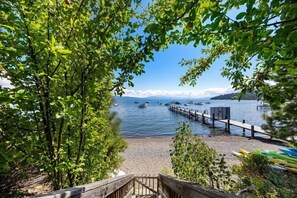 Eine Holztreppe führt zu einem Strand mit Booten im Wasser und einem Steg, der sich in den See erstreckt.