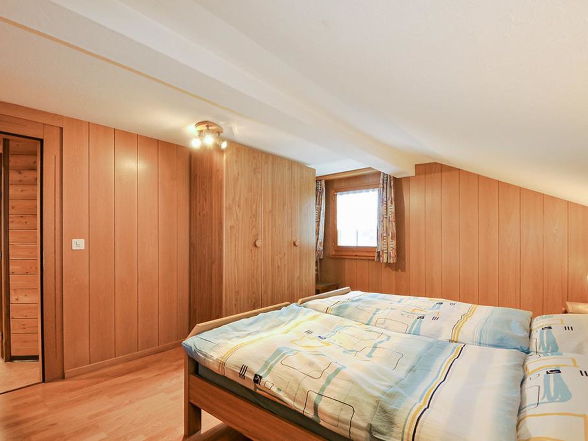 A bedroom with wooden paneling, a bed with patterned bedding, a window with curtains, and a ceiling light fixture.