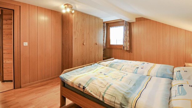 A bedroom with wooden paneling, a bed with patterned bedding, a window with curtains, and a ceiling light fixture. - Weissmies Appartement/Fewo 5-Bettwohnung by Interhome (Saas-Grund)