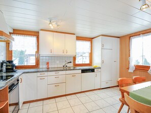 A kitchen with white cabinets, a countertop, a sink, and a dining area with a table and chairs. - Weissmies Appartement/Fewo 6-Bettwohnung by Interhome (Saas-Grund)