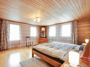 A bedroom with a wooden ceiling, a large bed, a dresser, and two windows with curtains. - Weissmies Appartement/Fewo 6-Bettwohnung by Interhome (Saas-Grund)
