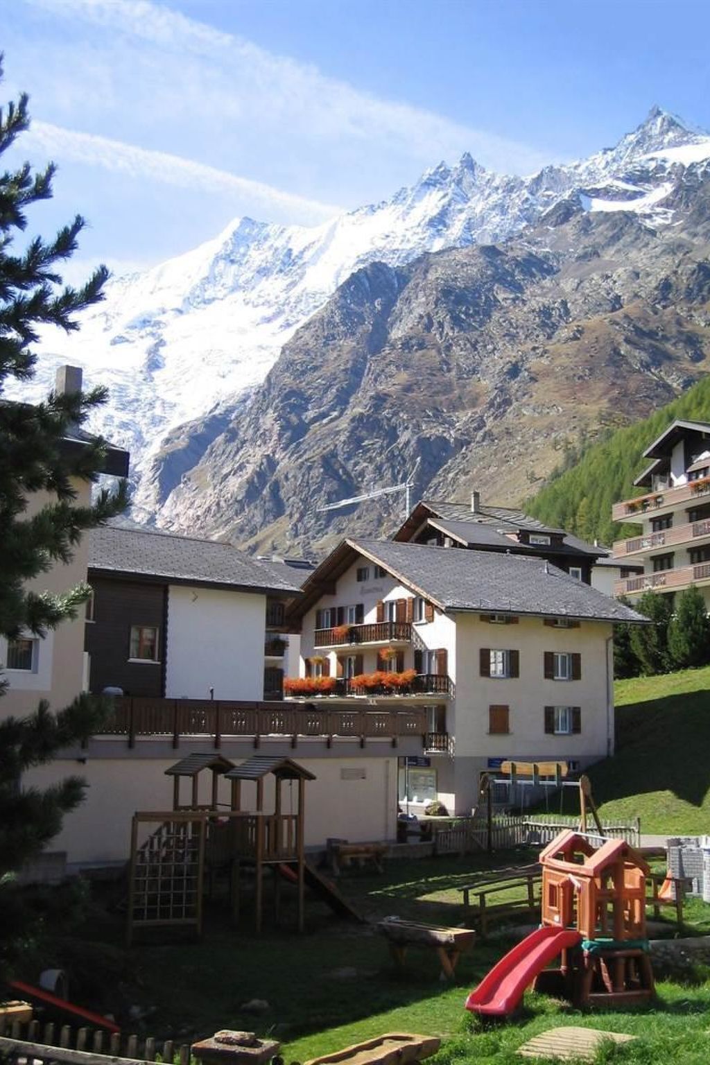 Un refugio de montaña con un parque infantil y un telesilla en primer plano, y montañas nevadas al fondo.