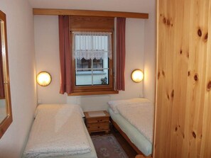 Ein kleines Zimmer mit zwei Einzelbetten, einem fensterbelaubten Fenster und Wandleuchten.