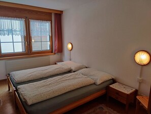 A bedroom with two beds, a window with curtains, a bedside table with a lamp, and a small chair. - Amethyst Appartement/Fewo 4-Bettwohnung Nr. 2 by Interhome (Saas-Fee)