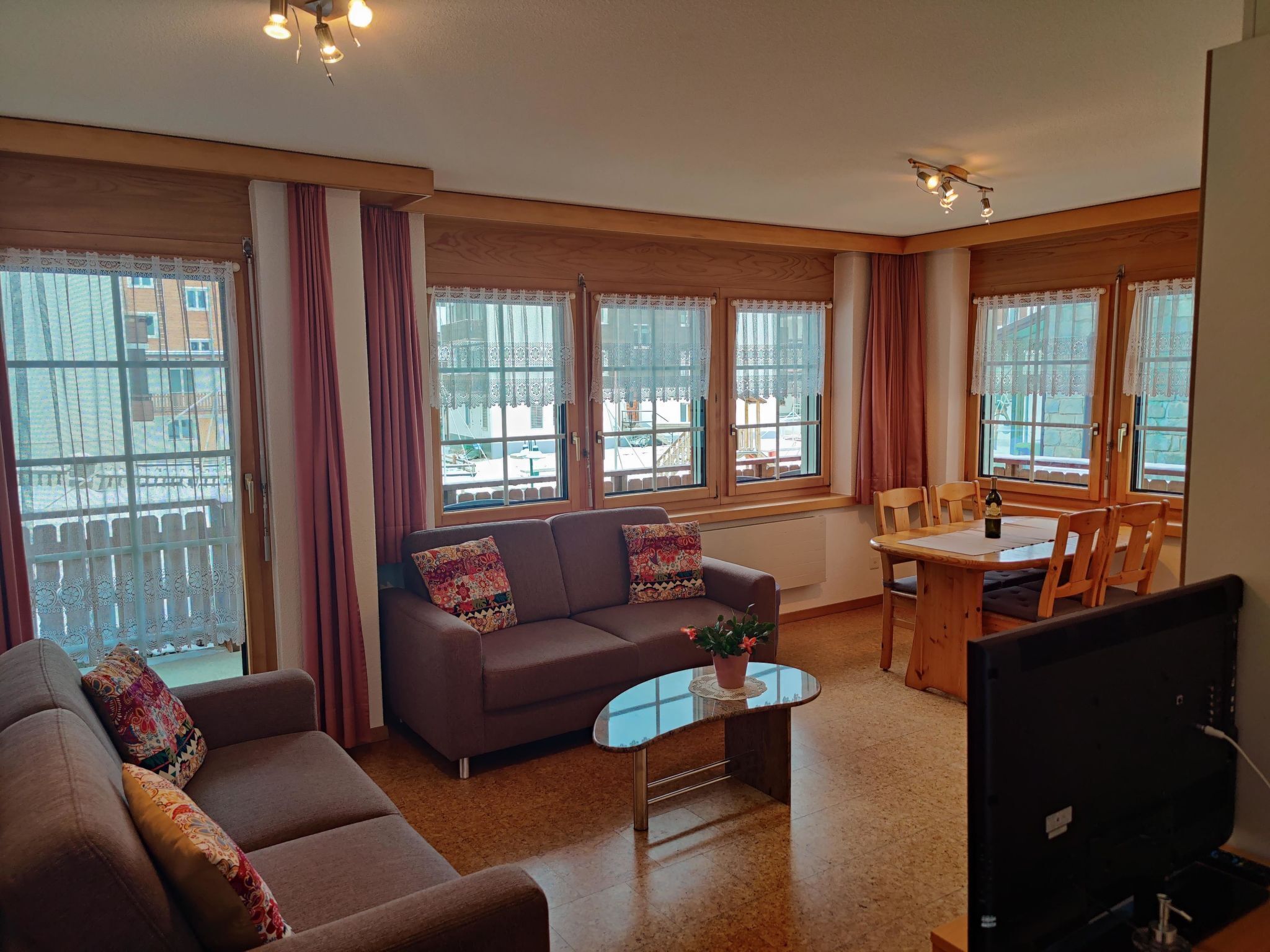 Una sala de estar con un sofá, una mesa de centro y un televisor. Hay ventanas con cortinas y vistas a edificios.