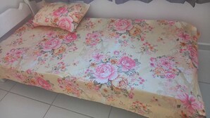 A bed with a floral bedspread and a matching pillow in a room with a plain wall and a window with curtains.