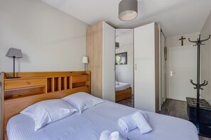 Ein Schlafzimmer mit hölzernem Kopfteil, einem Bett mit weißen Leinentüchern, einer Nachttischlampe, einem Kleiderschrank, einem Kleiderständer und einer Tür mit Kreuz.